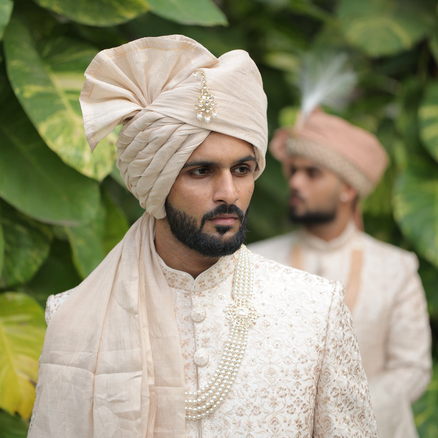 Man in traditional attire with a turban against a green leafy background