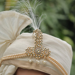 Beige turban with gold embellishments and a feather against a blurred green background