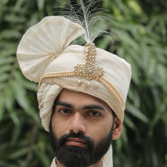 Man wearing a traditional cream-colored turban with a feather against a green leafy background