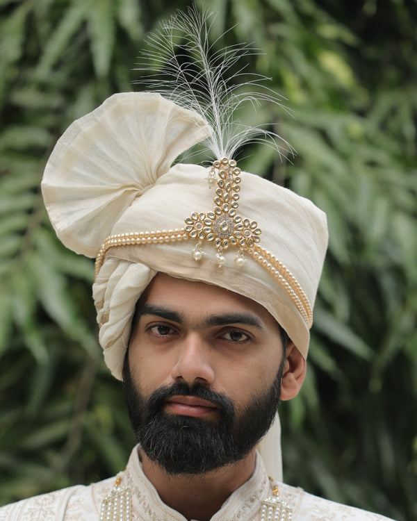 Man wearing a traditional cream-colored turban with a feather against a green leafy background