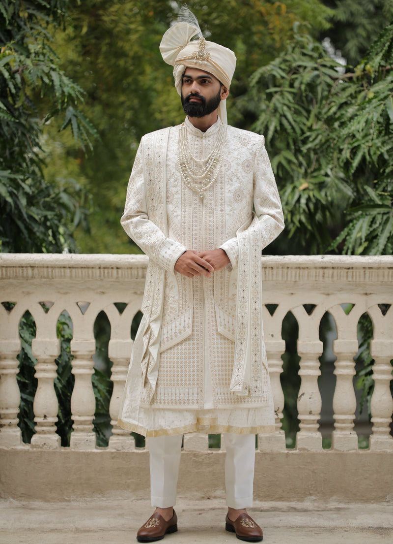 Man in traditional white outfit with a turban standing outdoors with greenery in the background